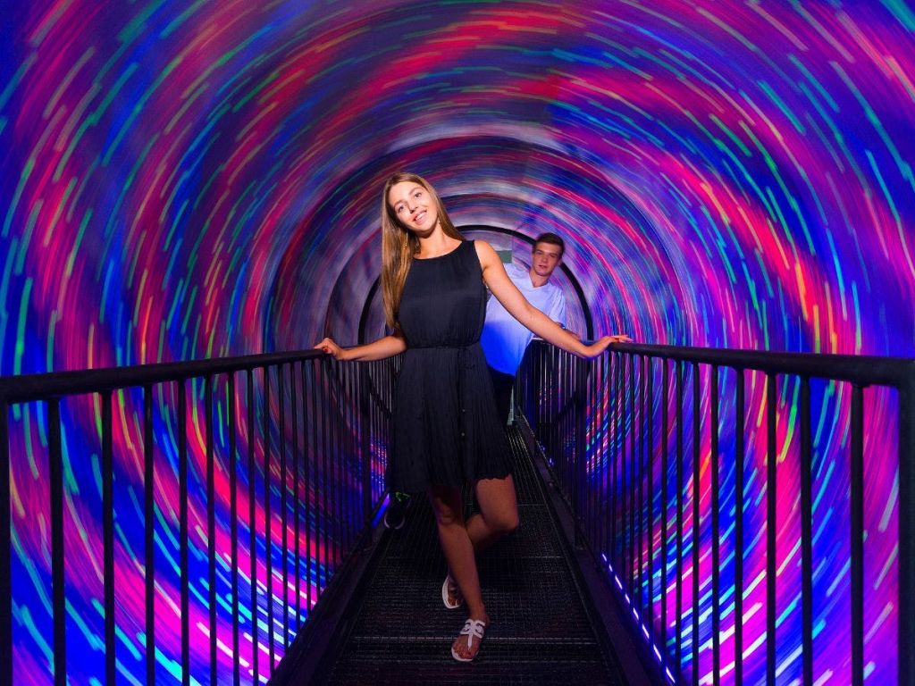 화려하고 소용돌이치는 LED 조명이 있는 회전하는 터널을 걷는 두 사람. 앞에 있는 여성은 둘 다 미소를 짓고 있습니다.