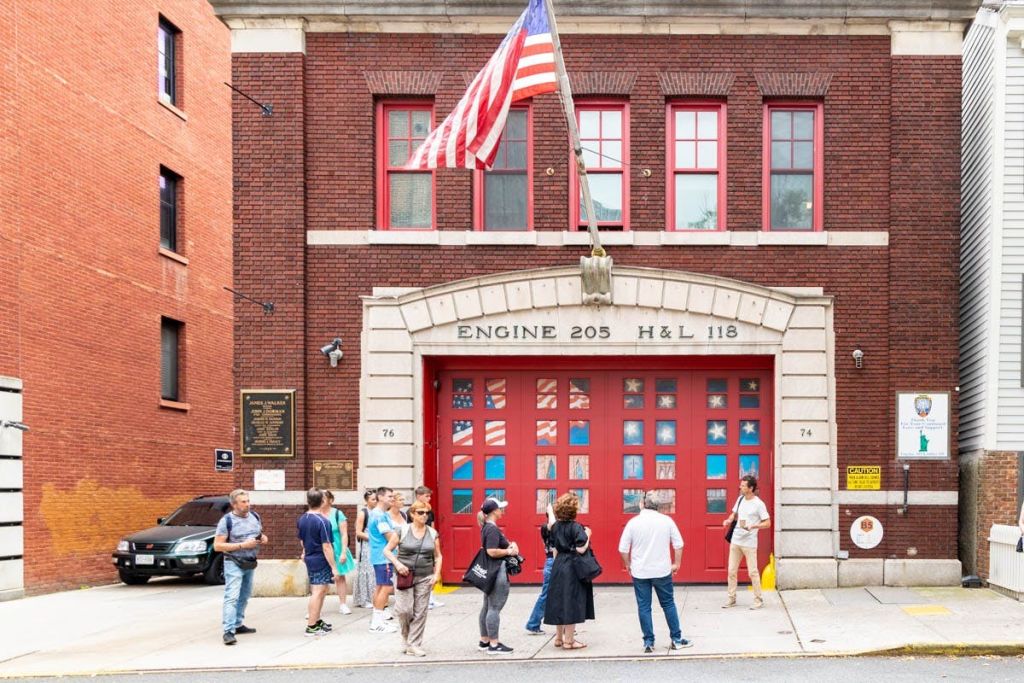 빨간색 문과 미국 국기가 날아다니는 소방서 앞에 서 있는 사람들. 건물에는 "엔진 205 H&L 118" 이라고 표시되어 있습니다.