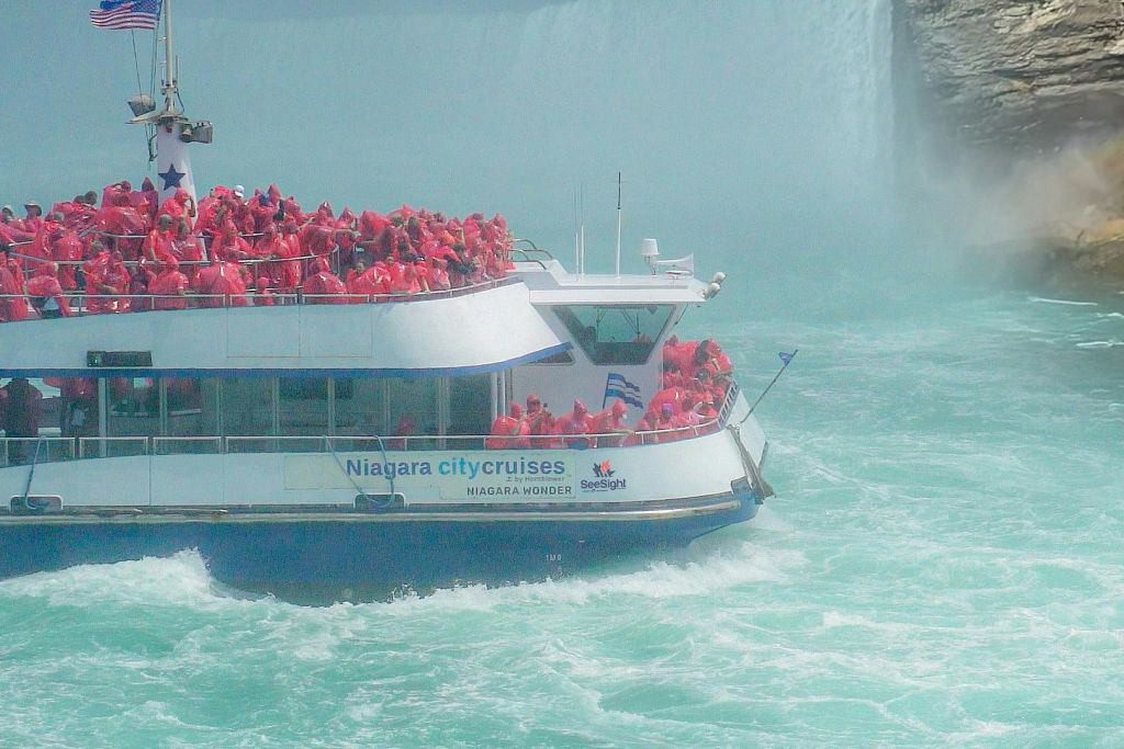 빨간색 판초를 입은 사람들로 가득 찬 보트가 "나이아가라 시티 크루즈" 라고 표시된 나이아가라 폴스로 다가갑니다.