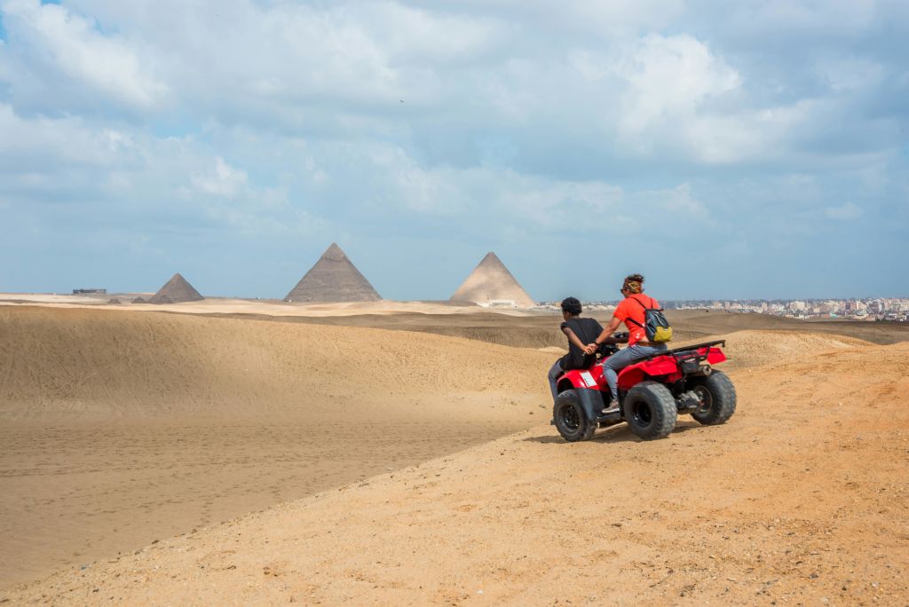 부분적으로 흐린 하늘 아래 피라미드를 배경으로 모래 사막에서 ATV를 타고 있는 두 사람.