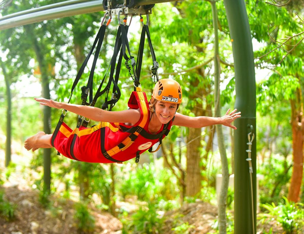주황색 헬멧을 착용한 빨간색과 노란색 안전 장비를 착용한 사람이 울창한 녹색 야외 환경에서 집라인 하네스에 매달린 채 미소를 짓고 있습니다.