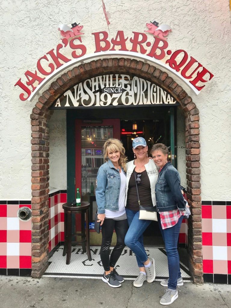 세 명의 여성이 레스토랑 입구 앞에서 'Jack's Bar-B-Que' 라고 적힌 간판을 들고 미소를 지으며 포즈를 취하고 있습니다.