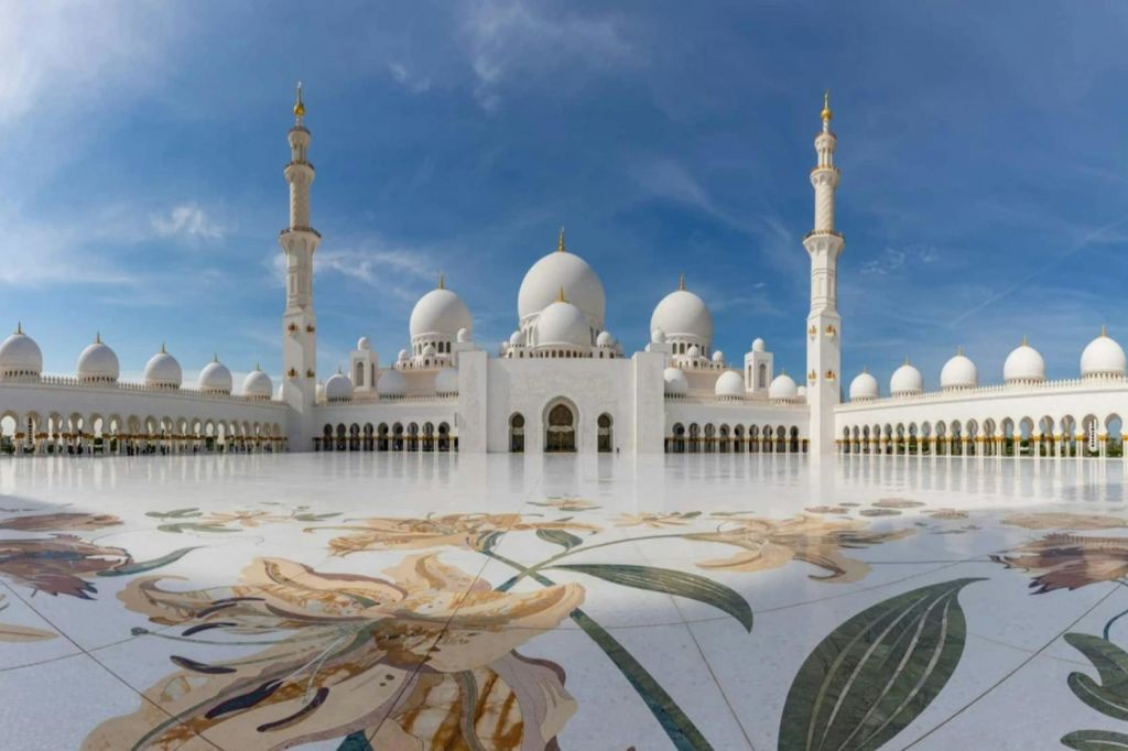 여러 개의 돔과 미나렛이 있는 흰색 모스크, 꽃 무늬가 있는 장식된 안뜰, 맑고 푸른 하늘 아래.