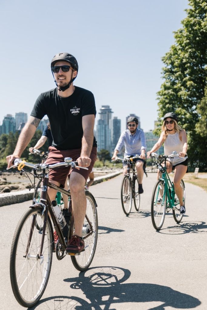 도시의 스카이라인과 커다란 나무를 배경으로 포장된 길에서 세 사람이 자전거를 타고 있습니다.