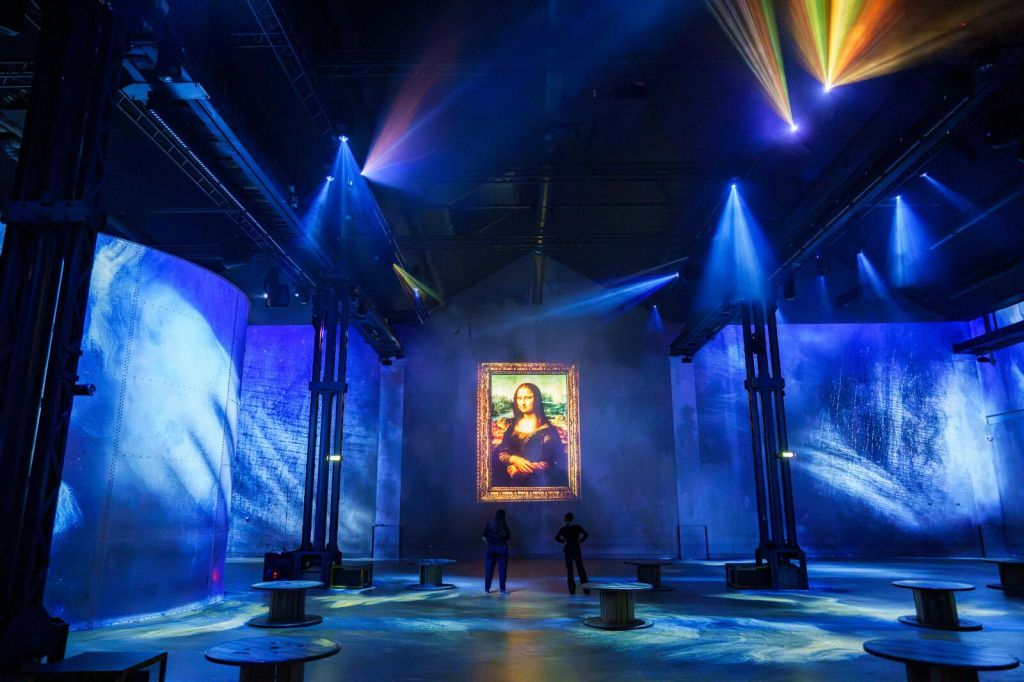 어두운 방에 모나리자 그림을 투영하여 화려한 불빛을 비추고 두 사람이 관찰하는 디지털 전시물입니다.
