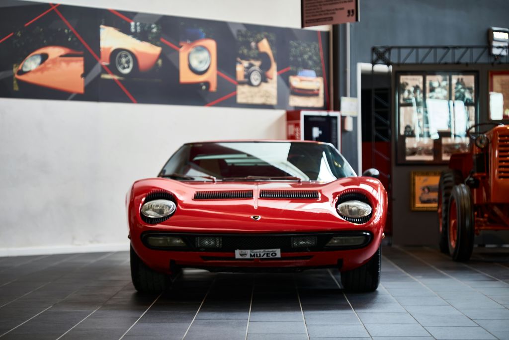 빨간색 클래식 스포츠카는 현대적인 박물관 환경에 전시되어 있으며, 뒤쪽 벽에 빈티지 자동차 사진이 있습니다.