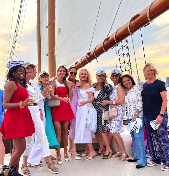 석양을 배경으로 커다란 돛이 달린 요트 데크에서 함께 포즈를 취하는 여성 그룹.
