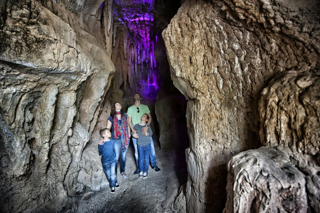 4인 가족이 보라색 빛으로 빛나는 바위 동굴을 탐험하며 위의 종유석을 올려다봅니다.