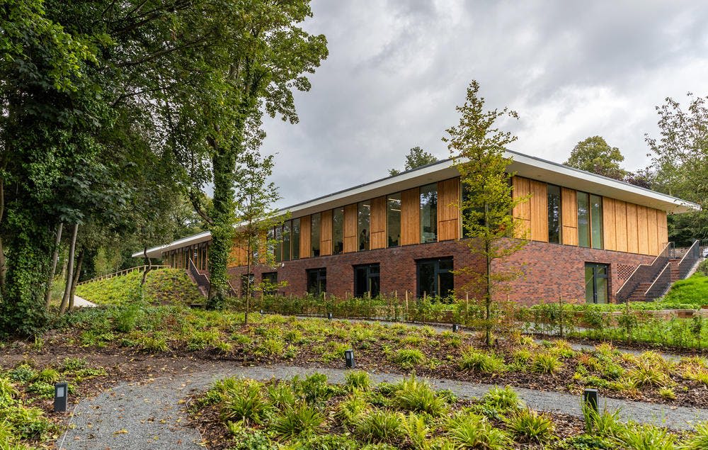 울창한 녹지와 자갈길로 둘러싸인 커다란 창문, 나무 패널, 벽돌 외관을 갖춘 현대적인 단층 건물입니다.