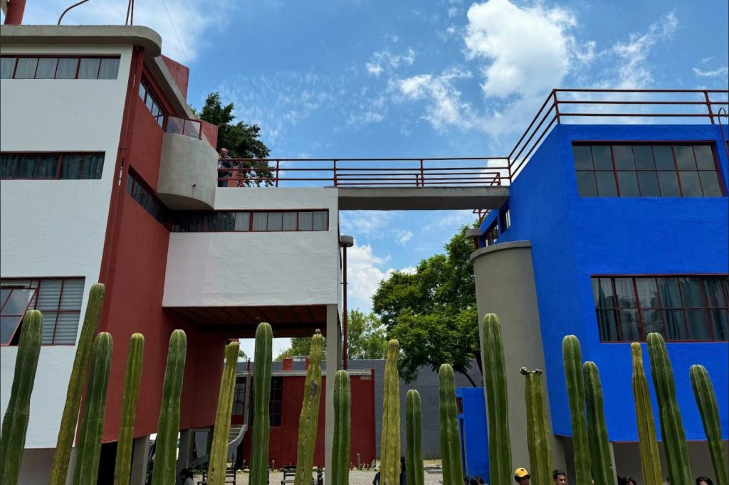 빨간색과 파란색의 현대적인 건물은 높은 통로로 연결되어 있으며, 앞쪽에는 높은 선인장이 있고 위쪽에는 부분적으로 흐린 하늘이 있습니다.