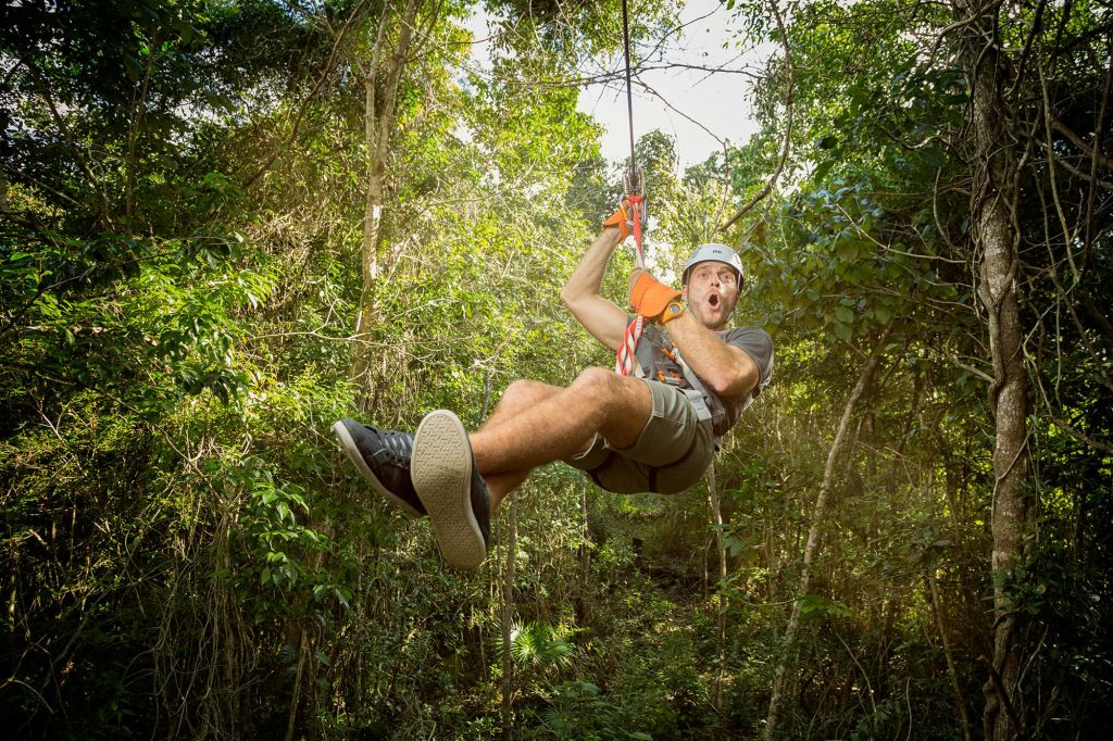 헬멧, 장갑, 캐주얼한 옷을 입고 흥분한 표정으로 빽빽한 숲을 가로지르는 집라인.