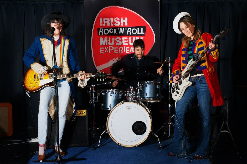 화려한 밴드 스타일의 복장을 한 사람이 드러머 옆에서 화이트 베이스 기타를 연주하고 뒤에 "Irish Rock 'N' Roll Museum Experience" 배너가 있습니다.