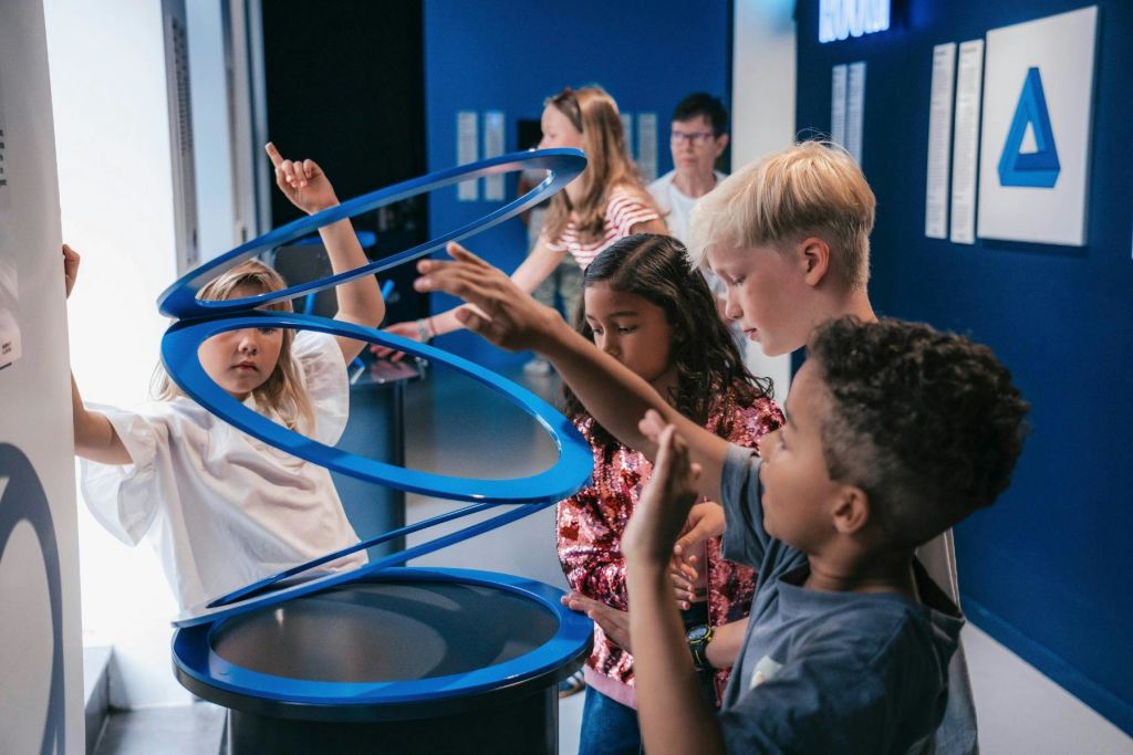 과학 박물관에서 파란색 나선형 전시물과 상호 작용하는 어린이들이 구조물에 참여하고 가리키고 있습니다.