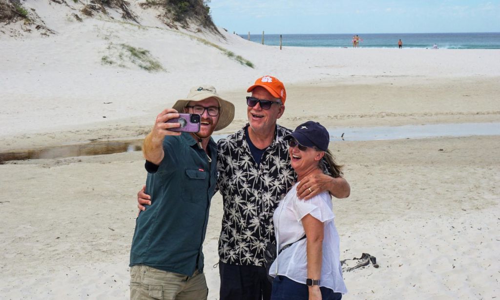 모래 언덕과 바다를 배경으로 모래사장에서 세 사람이 미소를 짓고 셀카를 찍고 있습니다.