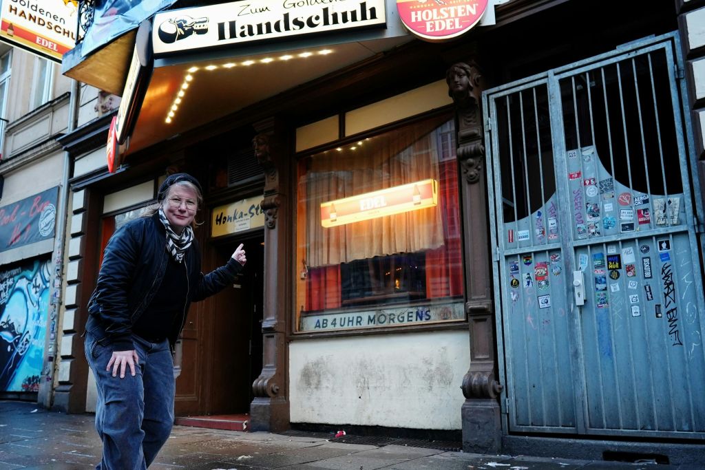 한 사람이 "Zum Goldenen Handschuh" 라고 적힌 표지판이 있는 길거리 사업장 밖에 서 있습니다. 입구를 가리키고 있습니다.