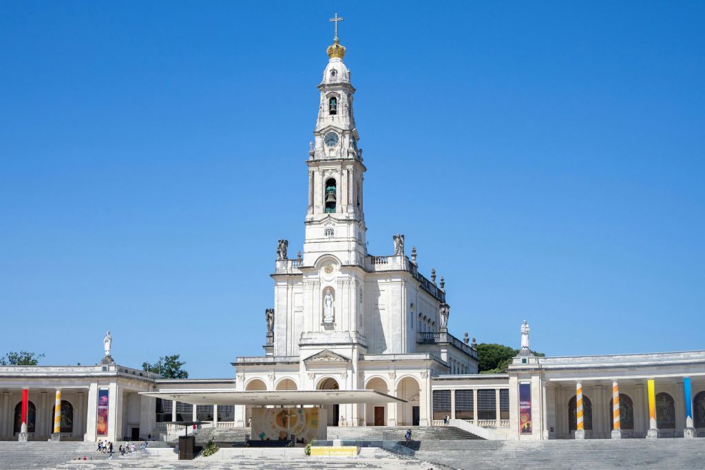 높은 종탑, 조각상, 아치형 창문, 파란 하늘을 배경으로 한 금색 십자가가 있는 흰색 바실리카의 전면 전망.