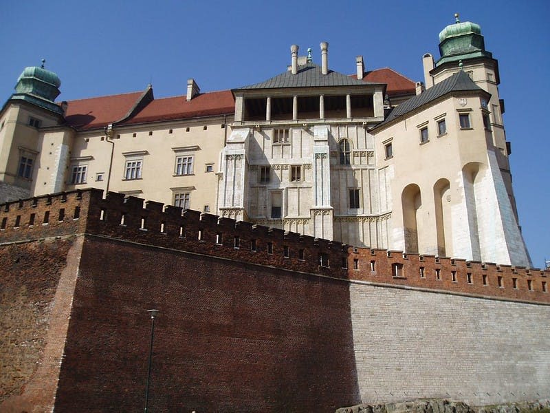 맑고 푸른 하늘 아래 높고 벽돌로 된 옹벽 뒤에 붉은 지붕과 흰색 벽이 있는 크고 유서 깊은 건물입니다.