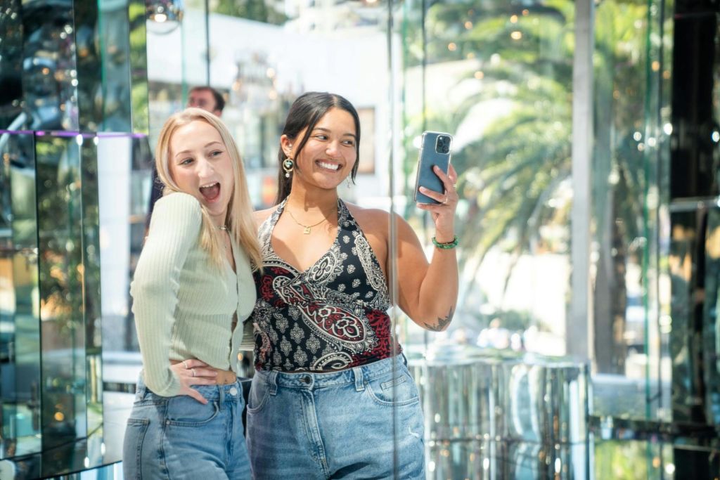 반사와 녹지를 배경으로 밝게 빛나는 유리 벽으로 꾸며진 방에서 함께 셀카를 찍는 두 여성.