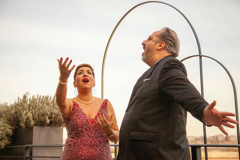 두 사람이 열정적으로 공연하고 있습니다. 한 사람은 빨간색 드레스를 입고, 다른 한 사람은 정장을 입고 야외를 배경으로 공연합니다.