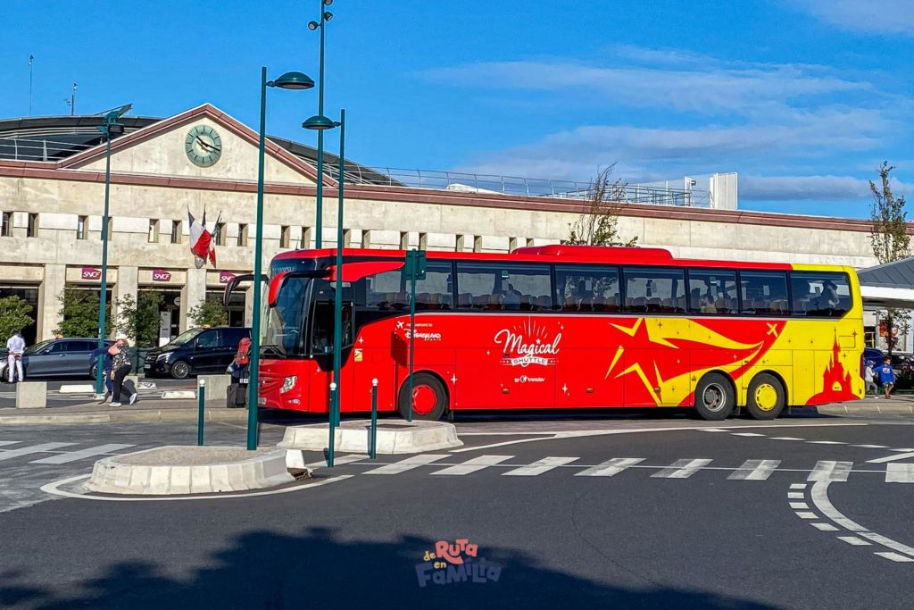 노란색 별 장식이 있는 빨간색 버스가 시계와 깃발이 있는 건물 근처 거리에 정차합니다.