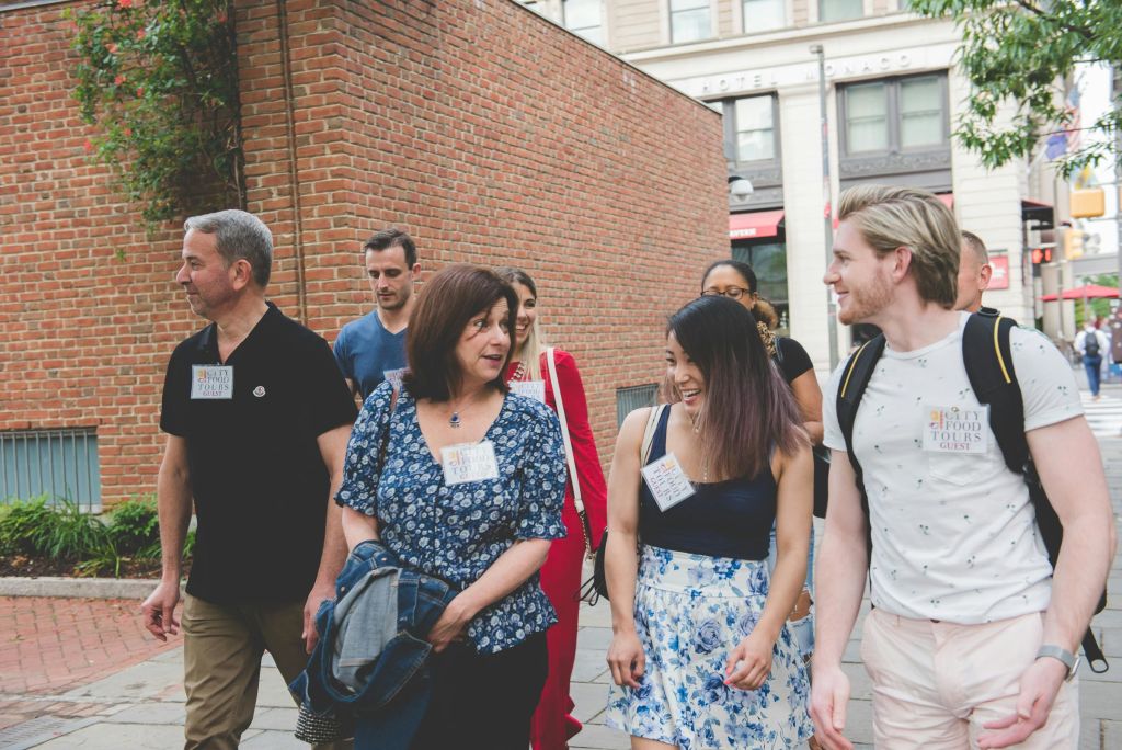 이름표가 붙은 사람들이 붉은 벽돌 벽에 둘러싸인 벽돌 포장된 거리를 걸으며 이야기를 나누고 있습니다.