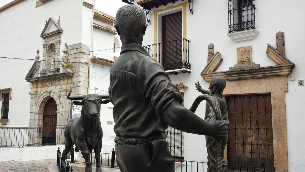 황소와 두 남자의 청동 조각상. 한 남자가 밧줄을 잡고 있습니다. 발코니가 있는 흰색 건물 앞에 있습니다.