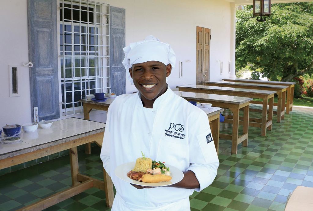 흰색 유니폼과 모자를 쓴 셰프가 나무 테이블, 타일 바닥, 커다란 창문이 있는 방에 서서 도금 식사를 하고 있습니다.