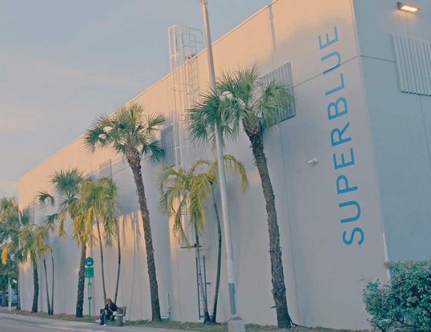 한 사람이 커다란 하얀 건물 옆 벤치에 앉아 있습니다. "SUPERBLUE" 라고 쓰여 있고, 높은 야자수가 줄지어 있습니다.
