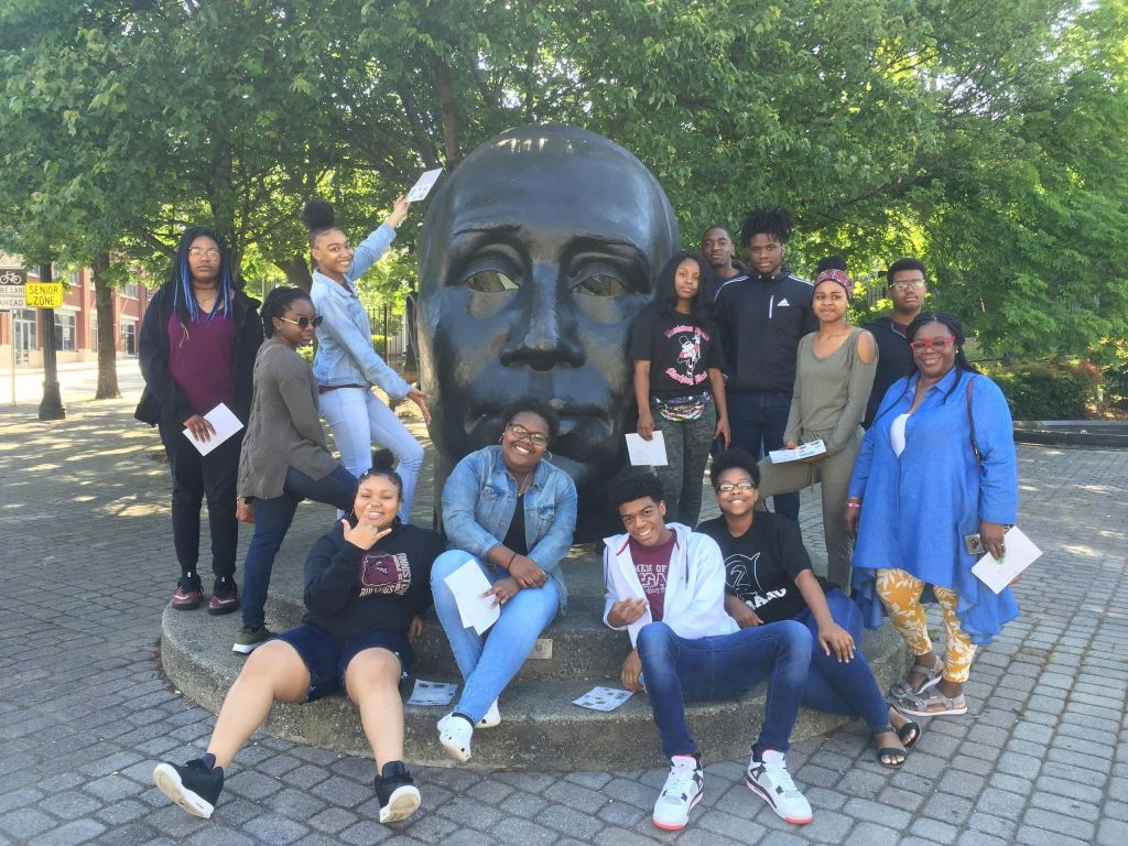 나무를 배경으로 공원의 커다란 머리 동상 앞에서 포즈를 취하고 있는 한 무리의 사람들.