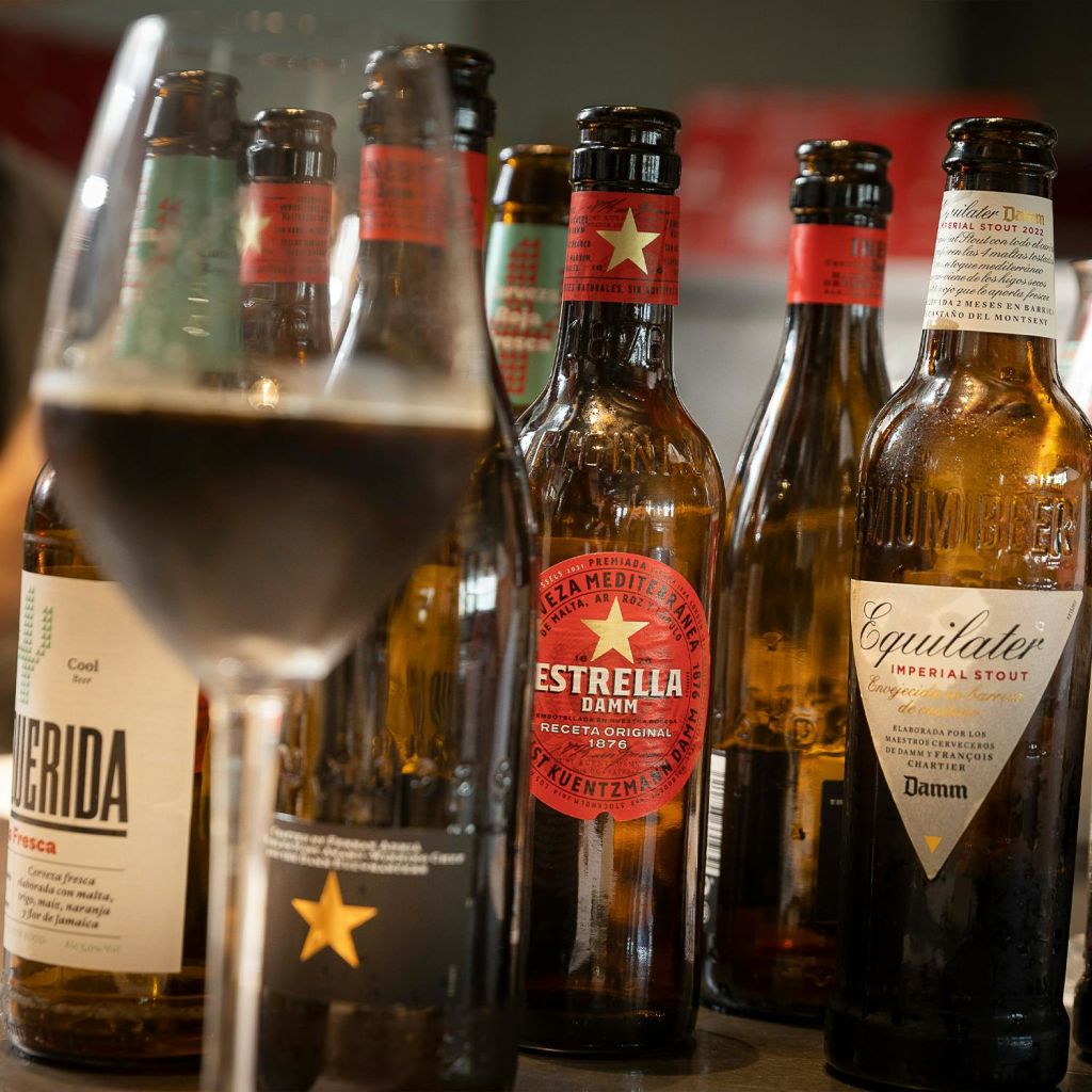 에스트렐라 댐 (Estrella Damm) 과 이퀼레이터 (Equilater) 등 다양한 라벨이 부착된 맥주병 몇 개가 전경의 전체 유리 옆에 놓여 있습니다.