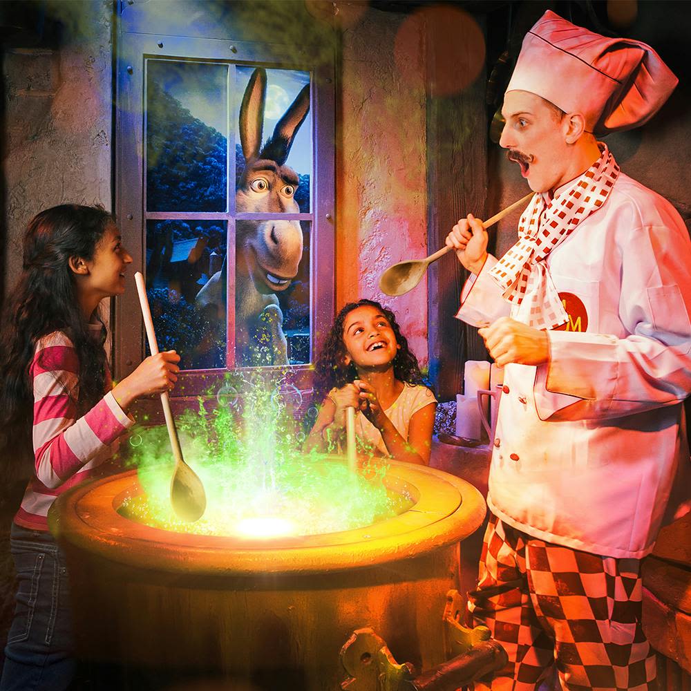 두 명의 소녀와 셰프가 창문 너머로 보이는 당나귀와 함께 빛나는 녹색 가마솥을 저어줍니다.