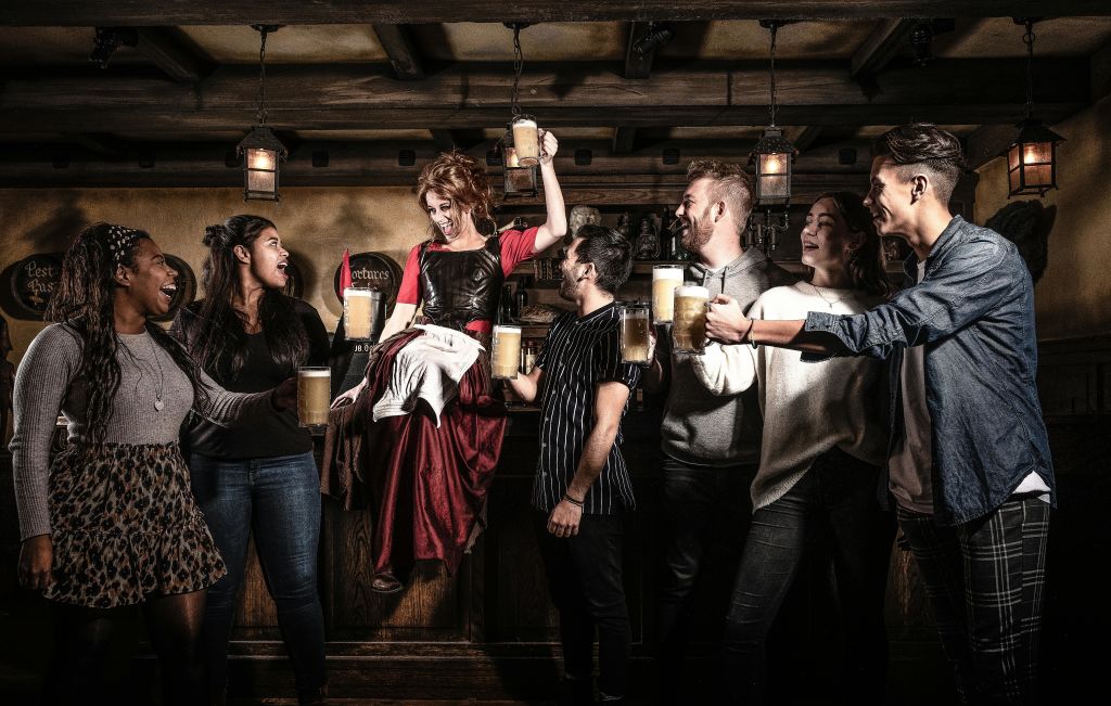 맥주 머그잔으로 건배하는 펍에서 활기찬 무리의 사람들, 중세 복장을 한 여성이 테이블에 서서 음료를 올리고 있습니다.