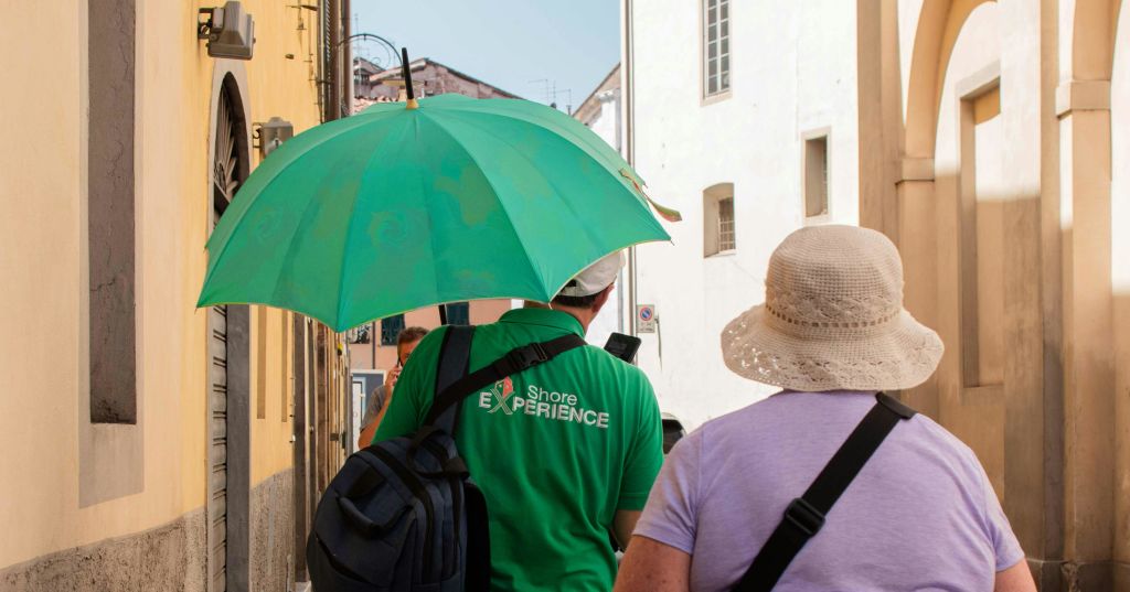 햇살 가득한 거리를 걷는 두 사람, 한 사람은 커다란 초록색 우산을 들고 '해안 체험' 이라고 적힌 초록색 셔츠를 입고 있습니다.