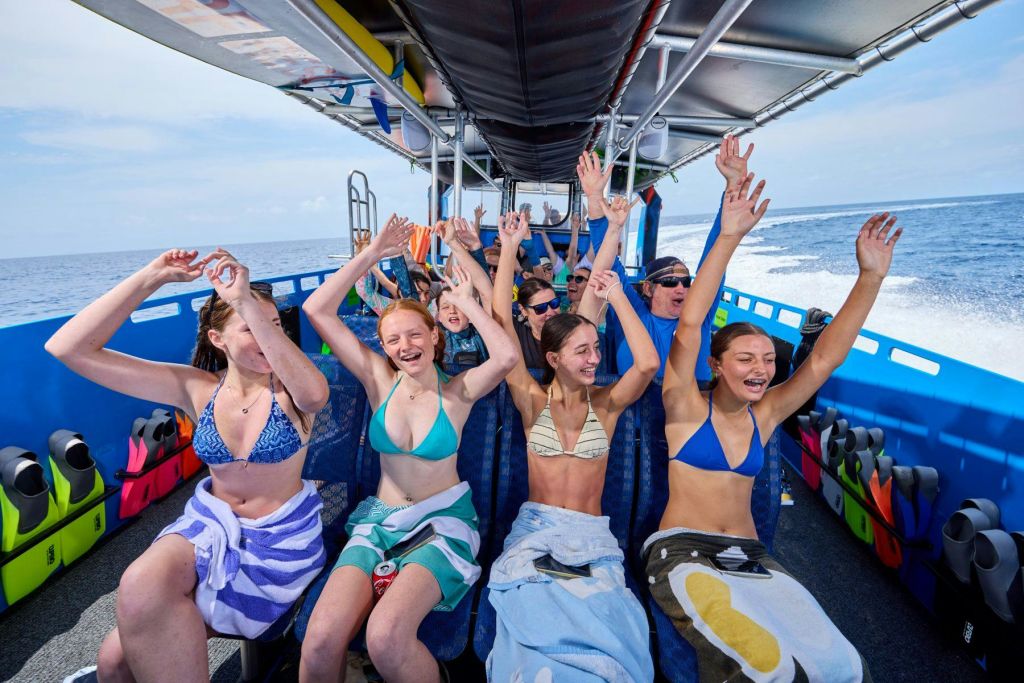 수영복과 수건을 입은 한 무리의 사람들이 보트를 타고 미소를 지으며 손을 들어올리고 있습니다.