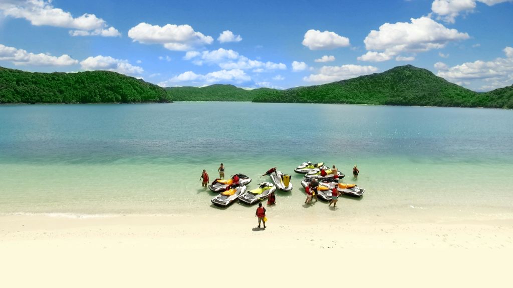 수영복을 입은 사람들은 푸른 언덕으로 둘러싸인 차분하고 투명한 호수 옆 모래 해변에서 제트 스키를 타고 모여듭니다.