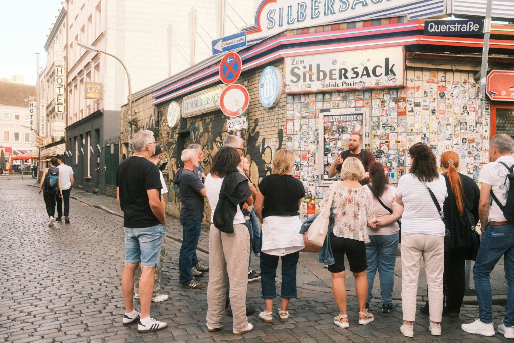 한 무리의 사람들이 스티커로 덮인 바 앞에 모여 "Zum Silbersack" 이라는 팻말을 들고 있습니다. 이 장면은 자갈길에 펼쳐집니다.