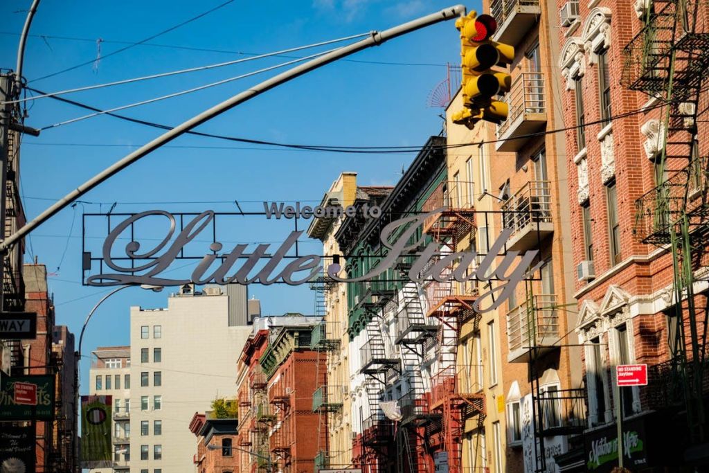 건물 사이에 '웰컴 투 리틀 이탈리아 (Welcome to Little Italy)' 라는 문구가 있고, 파란 하늘 아래 신호등과 불길이 흩날립니다.