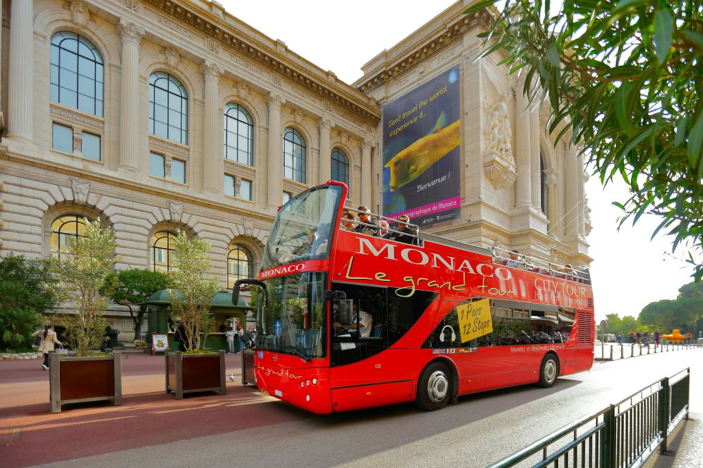아치형 창문이 있는 대형 클래식 건물 앞에 "모나코 르 그랜드 투어" 라고 표시된 빨간색 2층 투어 버스가 있습니다.