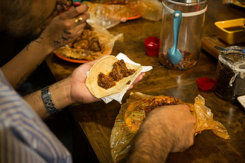 나무 테이블에 고기와 소스를 곁들인 타코를 준비하는 두 사람. 음식 용기와 접시가 놓여 있습니다.