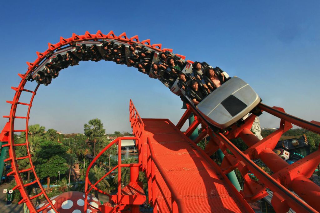 빨간색 롤러코스터를 탄 사람들이 맑고 푸른 하늘 아래 거꾸로 된 고리를 지나고 있습니다. 울창한 녹음이 아래에 보입니다.