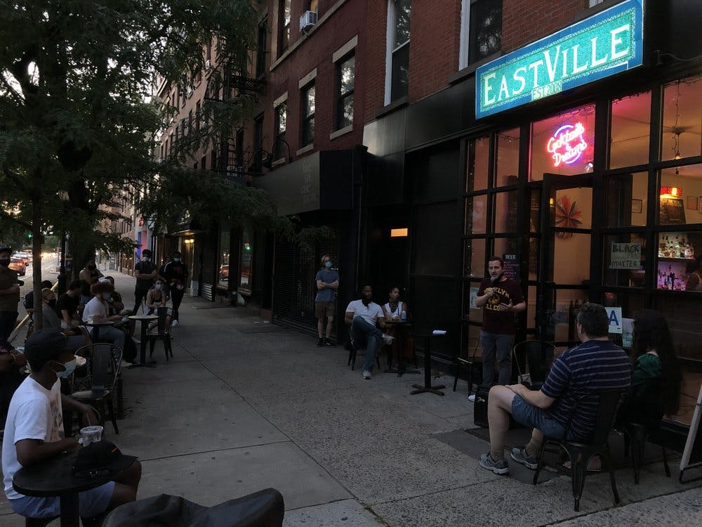 사람들이 이스트빌 코미디 클럽 바깥 해질 무렵 도시 보도에 앉아 서 있고, 일부는 공연자를 구경하고 있습니다.