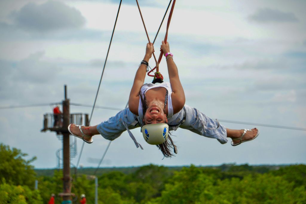 헬멧을 쓰고 지퍼라인을 거꾸로 누른 채 웃으며 팔과 다리를 벌리고 있는 사람. 초록색 나무가 배경입니다.