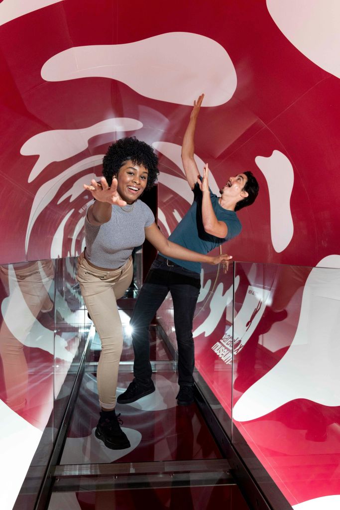 벽에 역동적인 패턴이 있는 빨간색과 흰색 터널 안에서 장난스럽게 포즈를 취하는 두 사람.