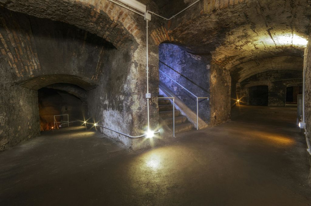 돌담, 아치형 출입구, 작은 조명으로 비춰지는 금속 난간 계단이 있는 희미한 지하 저장고.