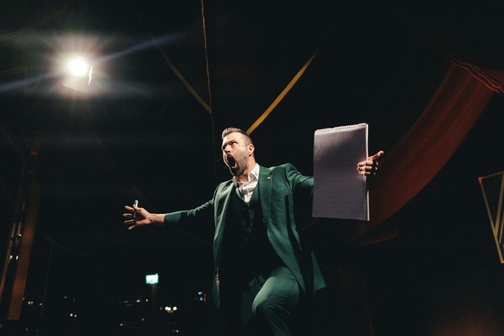 무대 조명 아래에서 메모장과 마커를 들고 열정적으로 말하는 녹색 양복을 입은 사람.