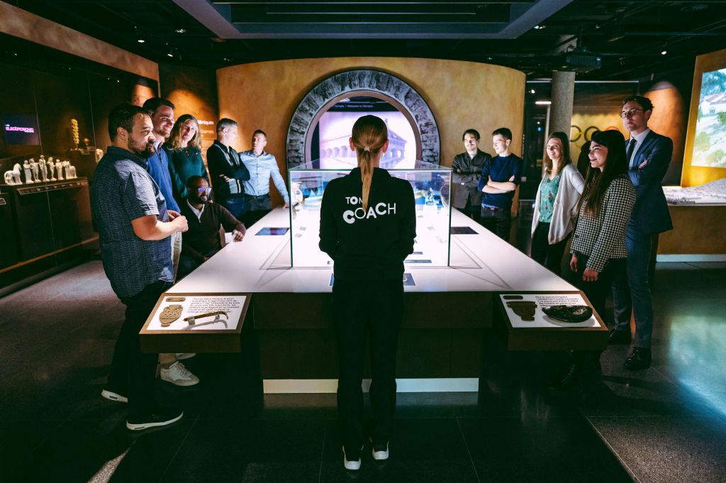 '코치' 라고 표시된 사람이 디스플레이 케이스 앞에 서서 현대적인 박물관 환경에서 한 무리의 사람들에게 프레젠테이션을 하고 있습니다.