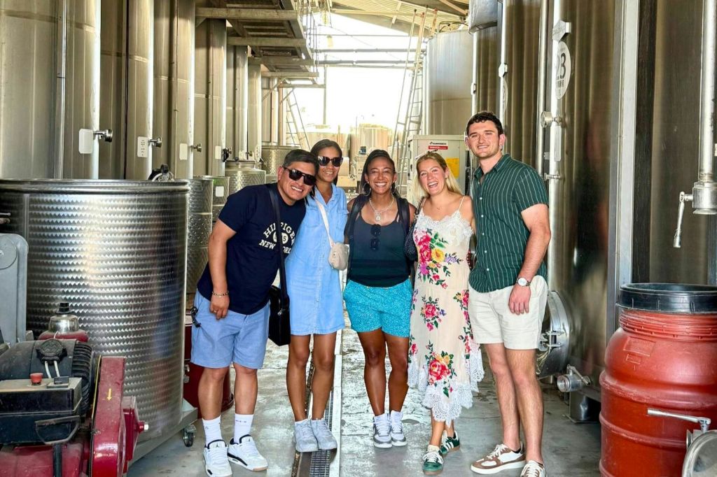 커다란 탱크와 밝은 조명을 배경으로 한 산업 시설에서 다섯 명이 함께 웃고 있습니다.