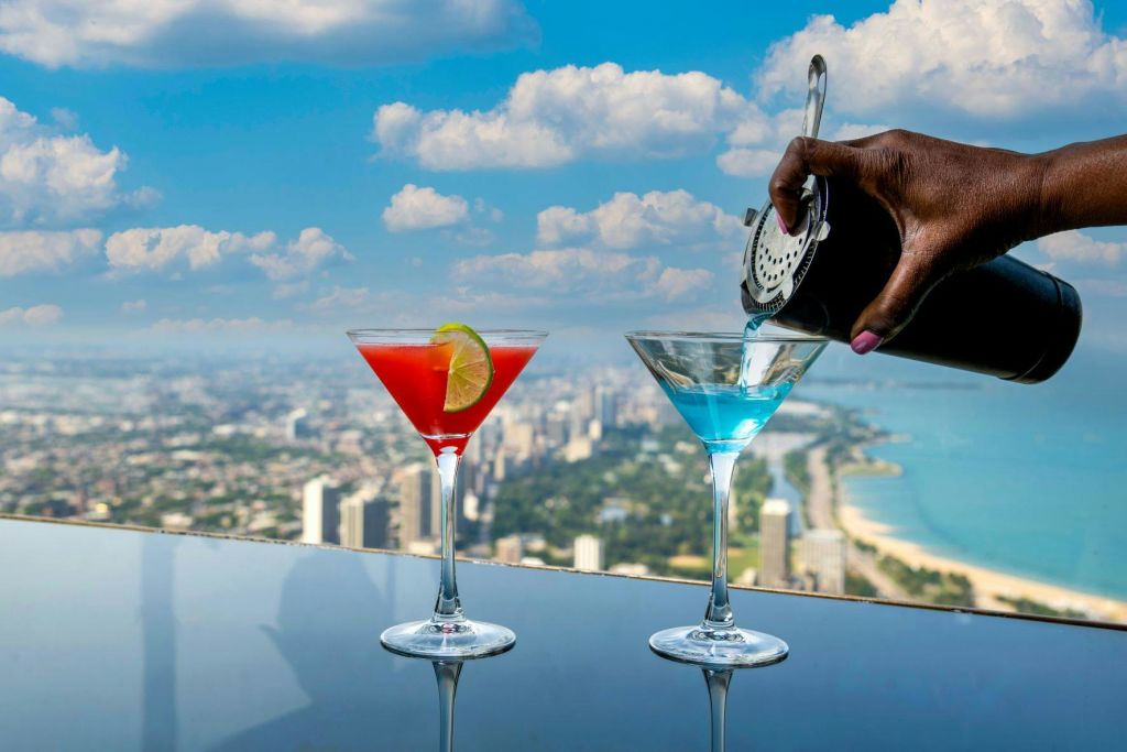 도시 풍경과 흐린 하늘을 배경으로 두 개의 칵테일, 라임 슬라이스가 있는 빨간색과 파란색. 셰이커에서 한 손이 음료를 쏟아냅니다.
