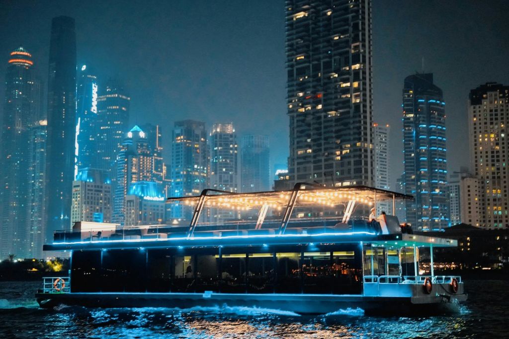 야간에 조명이 켜진 고층 빌딩을 배경으로 물 위를 항해하는 현대적인 보트입니다.