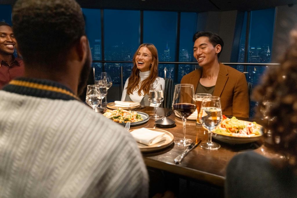 야간 도시 전망을 즐길 수 있는 레스토랑에서 저녁 식사를 즐기는 사람들, 음식과 음료가 있는 테이블에서 웃으며 대화하는 사람들.
